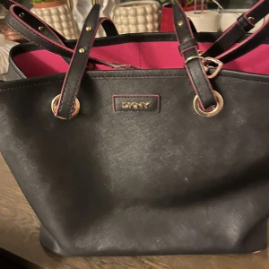 Svart handväska från DKNY med rosa insida - Snygg svart handväska från DKNY med rosa insida och rosa detaljer längs kanterna. Väskan har guldfärgade metalldetaljer och flera remmar med justerbara spännen. Materialet är skinn med en lätt textur. Perfekt för dig som gillar stilrena och färgglada accessoarer.