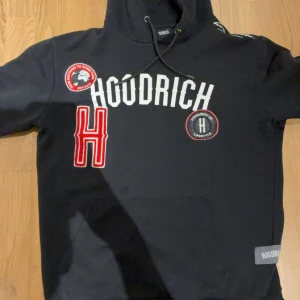 Svart hoodie från Hoodrich med patchar - Svart hoodie från Hoodrich med stora vita och röda tryck och patchar framtill. Klassisk huva med snörning och känguruficka. Snygga detaljer med broderade märken och stor röd H på bröstet. Perfekt streetwear-vibe.