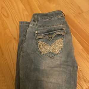 Blå jeans med broderade vingar - Snygga ljusblå jeans med unika broderade vingar och paljettdetaljer på bakfickan. Jeansen har klassisk femficksdesign och en cool spetsdetalj på ficklocket. Perfekta för dig som vill sticka ut med din stil.