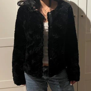 Svart pälsjacka i mjukt material - Mysig svart pälsjacka med rund halsringning och knäppning framtill. Jackan har lång ärm och en rak, något loose passform. Perfekt för dig som vill ha en stilren och fluffig look. Tyvärr har en knapp ramlar ut men det är verkligen inget som syns.