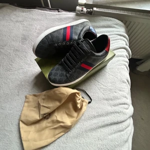 Gucci sneakers med röd/blå rand - Säljer ett par snygga Gucci sneakers i mörkgrått med GG-monogram, röd och marinblå rand på sidan och röd detalj vid hälen. Skorna har vita sulor, snörning och är tillverkade i canvas och skinn. Perfekta för dig som vill sticka ut med lyxig streetstyle. Dustbag och låda ingår. 