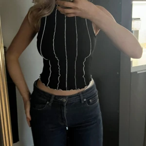 Svart croppad linnetopp från NA-KD - Säljer en svart croppad linnetopp från NA-KD med vita vågiga vertikala sömmar. Toppen är ärmlös, ribbad och har hög hals. Perfekt för dig som gillar trendiga detaljer och en tight passform.