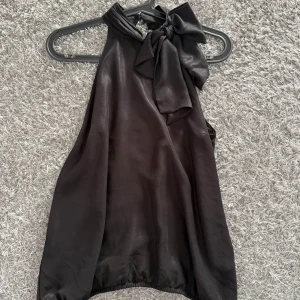 Svart halterneck blus med rosett - Snygg svart halterneck blus i glansigt material med stor rosett vid halsen och öppen rygg. Blusen har en lös passform och resår nertill för en cool siluett. Perfekt för dig som vill sticka ut med en elegant och trendig look.