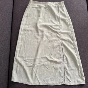 Ljusgrön blommig midikjol Vero Moda - Superfin ljusgrön kjol från Vero Moda med diskret blommigt mönster och slits på sidan. Kjolen har en rak passform och resår i midjan för extra komfort. Perfekt för dig som gillar en stilren och feminin look. Kjolen är ny och är oanvänd.