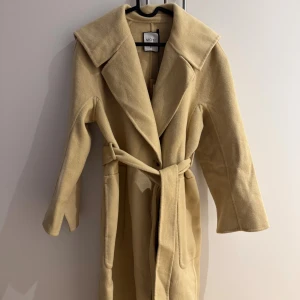 Beige/ gul kappa från MOTF med bälte XS - Säljer en gul kappa från MOTF i storlek XS. Kappan har bred krage, knytskärp i midjan och långa ärmar. Tillverkad i mjukt material som känns lyxigt och passar perfekt till höst och vinter. Enkel och stilren design med fickor på sidorna. Kappam är hel ny och har aldrig använt.