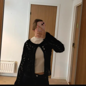 Svart stickad kofta från Jinska - Säljer en svart stickad kofta från Jinska i storlek S. Koftan har ett öppet, luftigt mönster och långa ärmar. Perfekt att slänga över en topp för en chill och snygg look. Supermjuk och lätt att styla till jeans eller kjol.