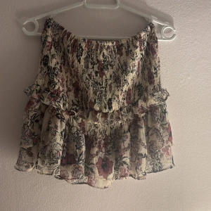 Blommig volang kjol  - Supersöt populär volangkjol med blommigt mönster i beige, vinrött och svart. Kjolen har volangdetaljer samt smockad struktur som ger en snygg passform. 