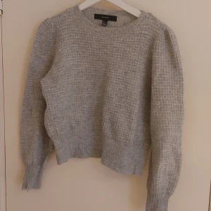 Grå stickad tröja från Vero Moda - Mysig grå stickad tröja från Vero Moda i storlek M. Tröjan har rund halsringning och långa ärmar med breda muddar. Tröjan har ett litet hål (se bild) och äe även lite nopprig. 