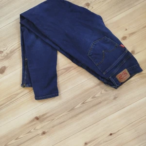 Levi's blå skinny jeans med dragkedja nedtill.  - Mörkblå skinny jeans från Levi's med klassiska detaljer och dragkedja vid benslut. Jeansen har fem fickor, Levi's patch bak och guldfärgade sömmar. Perfekta för dig som gillar en tajt passform och stilren look.