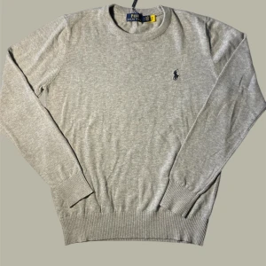 Grå stickad tröja från Polo Ralph Lauren - Snygg grå stickad tröja från Polo Ralph Lauren med klassisk rund hals och diskret broderad logga på bröstet. Tröjan har långa ärmar och ribbade muddar vid ärmslut och nederkant. Perfekt för dig som gillar stilrena och tidlösa plagg.
