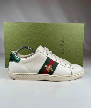 Gucci Ace sneakers med bi-detalj - Gucci Ace sneakers i vitt skinn med klassiska gröna och röda ränder på sidan, broderad guldfärgad bi och metallicgrön samt röd hälpanel. Rund tå, platt sula och snörning framtill. Snygg och ikonisk modell som sticker ut med sina lyxiga detaljer. Detta är en förbeställning, snabbasg frakt 3 dagar.