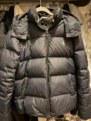 Mörkblå pufferjacka Calvin Klein  - Säljer en mörkblå pufferjacka från Calvin Klein Jeans med hög krage, dragkedja och logotyp på bröstet samt ett stort CK-märke på ärmen. Jackan har långa ärmar och praktiska fickor. Perfekt för kalla dagar och riktigt snygg stil.