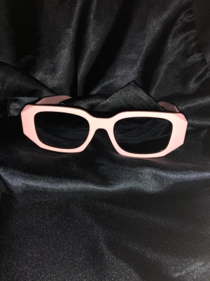 Rosa fyrkantiga solglasögon från Prada - Säljer ett par statement solglasögon från Prada med breda, fyrkantiga bågar i ljusrosa. Modellen är 8251 och tillverkad i Italien. Glasen är svarta och bågarna har en cool, chunky design med logga på sidan. Perfekt för dig som vill sticka ut med en trendig accessoar.