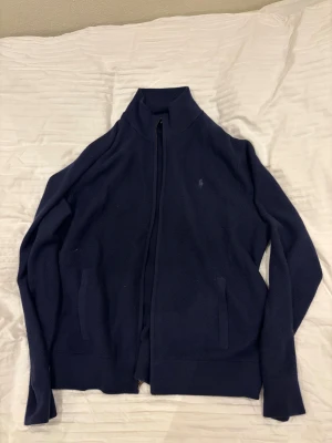 Marinblå XL kofta från Polo Ralph Lauren  - Snygg marinblå kofta från Polo Ralph Lauren i tvättbar merinoull. Tröjan har hel dragkedja framtill, ståkrage och två fickor. Diskret broderad logga på bröstet och ribbade muddar vid ärmslut och nederkant. Perfekt för dig som gillar klassisk stil. Storlek XL