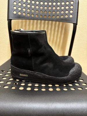 Svarta mockaboots från Bally - Säljer ett par svarta boots från Bally i mjuk mocka med grov gummisula och rund tå. Skorna har dragkedja baktill för enkel på- och avtagning. Diskret Bally-logga på sidan och sula. Perfekt för dig som gillar stilrena och robusta skor.