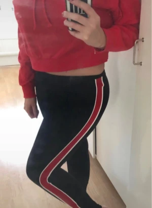 SET! Leggings med hoodie från H&M - Snygg röd croppad hoodie med huva och snörning framtill. Svarta leggings med en röd och vit rand längs utsidan av benen. De har en tight passform och hög midja, vilket ger en sportig och chill look. TRÖJAN ÄR I STORLEK M OCH BYXAN I STORLEK S