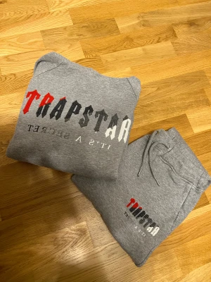 Grå mjukisbyxor Trapstar med tryck - Säljer ett par grå mjukisbyxor från Trapstar med coolt tryck i rött, svart och vitt på benet. Byxorna har snörning i midjan och är gjorda i mjukt bomullsmaterial. Perfekt för chill dagar och streetwear-stil.