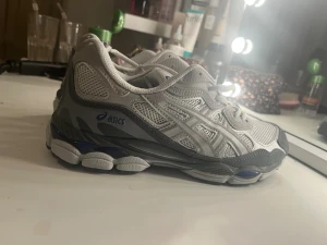 Asics GEL sneakers i grå och vit - Säljer ett par Asics GEL sneakers i grått och vitt med blå detaljer. Skorna har meshpaneler för extra ventilation, syntetiska överdrag och en robust, mönstrad sula för bra grepp. Klassisk snörning och rund tå. Perfekta för dig som gillar sportig stil.