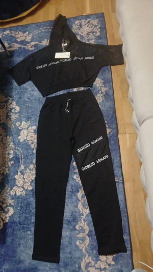 Svarta mjukisbyxor Giorgio Armani - Svarta mjukisbyxor från Giorgio Armani med tryckt logga på båda benen. Byxorna har elastisk midja med snörning och meshdetaljer på benen. Materialet är mjukt och bekvämt, perfekt för chill eller träning. Snyggt och stilrent set med matchande mesh-hoodie. Finns i andra storlekar och kom gärna och skriv ifall ni vill att jag mäter och kollar😇
