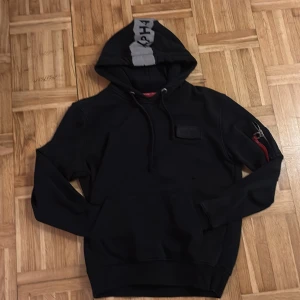 Svart hoodie Alpha Industries i Storlek S - Svart hoodie från Alpha Industries med huva och snörning, samt coolt märke på bröstet. Tröjan har en ficka på magen och en extra ficka med dragkedja på ärmen. Insidan är mjuk och skön, perfekt för chill dagar. Snygga detaljer på huvan och bröstet.