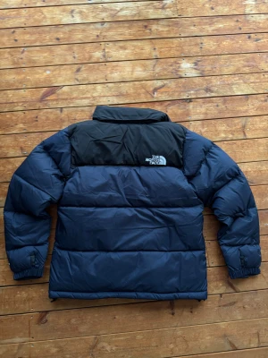 The North Face 1996 Retro Nuptse  - Blå/svart The North Face 1996 Retro Nuptse dunjacka i storlek S. Klassisk och varm vinterjacka med ikonisk boxig passform. Jackan är helt ny med prislapp kvar – perfekt för kalla dagar och tidlös i stilen.