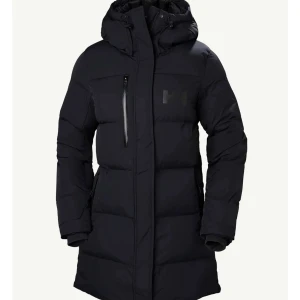 Svart pufferjacka från Helly Hansen - Svart pufferjacka från Helly Hansen med huva, dragkedjor och snörning i guld. Jackan har en quiltad design och flera praktiska fickor med dragkedja. Perfekt för kalla dagar och har en riktigt schysst streetstil. (Nypris 3699kr)