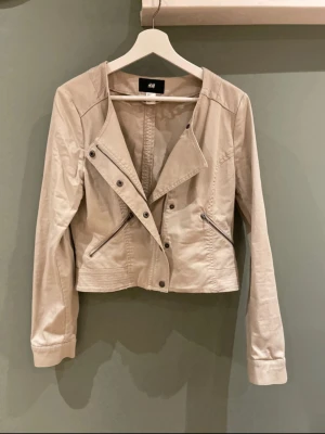 Beige bomberjacka från H&M - Snygg beige bomberjacka från H&M med rundad hals och coola detaljer som dragkedjor vid fickorna och knappar framtill. Jackan har lång ärm och är tillverkad i mjuk bomull, perfekt för dig som gillar stilrena och enkla plagg.