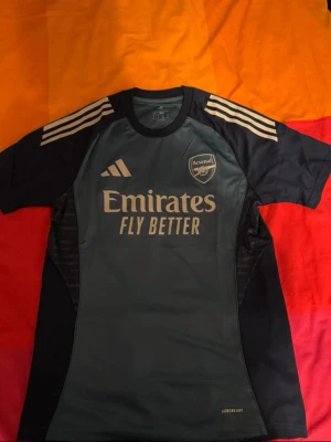 Arsenal tränings-tshirt Adidas M - Snygg Arsenal tränings-tshirt från Adidas i marinblått och mörkgrått med vita detaljer. Kortärmad modell med klassiska Adidas-ränder på axlarna och tryck med Emirates Fly Better på bröstet. Tillverkad i lätt och ventilerande polyester, perfekt för fotbollsträning.