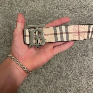 Burberry bälte med silverdetalj - Snyggt Burberry-bälte i klassiskt rutigt mönster med beige, svart och röda toner. Bältet har en bred design och en stor silvrig spänne med Burberrys ikoniska TB-logo. Tillverkat i skinn med präglade detaljer och typisk Burberry-stil.