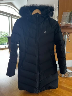 Svart lång dunjacka Calvin Klein Jeans - Svart lång dunjacka från Calvin Klein Jeans med fluffig huva och snyggt CK-logo på bröstet. Jackan har dragkedja framtill, två sidofickor med dragkedja och quiltad design. Perfekt för kalla dagar med sin varma och stilrena look.