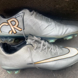 Nike CR7 silver fotbollsskor - Snygga silverfärgade Nike CR7 fotbollsskor med orange och svart detalj på sidan och CR7-logga. Skorna har snörning och platt sula med dobbar för bra grepp på planen. Ovandelen är i syntetmaterial och insidan är svart. Perfekta för dig som vill sticka ut på fotbollsplanen.