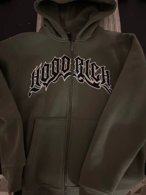 Olivgrön hoodie från Hoodrich - Säljer en olivgrön hoodie från Hoodrich i storlek S. Tröjan har dragkedja, huva med broderad text och stort Hoodrich-tryck i svart och vitt på bröstet. Mjuk bomullsmix, två fickor fram och ribbade muddar. Perfekt för streetwear-stilen.