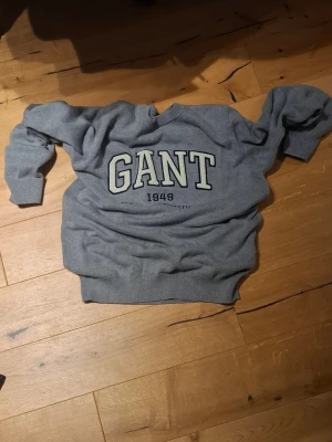 Grå sweatshirt från GANT - Säljer en grå sweatshirt från GANT med stor logotyp och texten '1949' på bröstet. Tröjan har rund hals, ribbade muddar vid ärmslut och nederkant samt långärmad passform. Perfekt för dig som gillar klassisk och sportig stil.