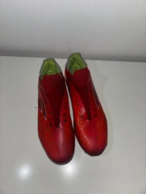 Röda Adidas fotbollsskor - Säljer ett par röda Adidas fotbollsskor med silverfärgade detaljer och grå häl. Skorna har en strömlinjeformad design utan synlig snörning och insida i limegrönt. Yttermaterialet är syntet och sulan har dobbar för gräsplan. Perfekt för dig som vill sticka ut på planen. Pris kan diskuteras 