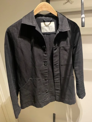 Svart overshirt från These Glory Days - Svart overshirt från These Glory Days med klassisk krage, tre framfickor och knäppning med svarta knappar. Jackan är tillverkad i ett slitstarkt bomullstyg och linne. Den har en stilren, rak passform som funkar till många olika looks.