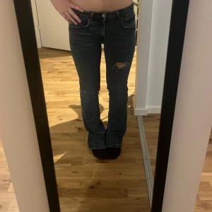 Bootcut jeans med slitningar - Säljer ett par mörkblå bootcut jeans med slitningar på knäna och klassiska fem fickor. Jeansen har låg midja och är tillverkade i ett robust denimtyg. Snygga kontrastsömmar och en cool, avslappnad vibe. Dom sitter konstigt på mig och ena benet rullar fram 