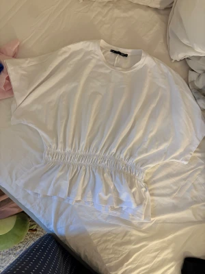 Vit och svart blus med volang - Två blusar från Lager 157 i storlek L, en vit och en svart. Båda har rund hals, korta ärmar och en snygg volangkant nedtill med smockad midja som ger en cool siluett. Perfekt för dig som gillar enkel men trendig stil.