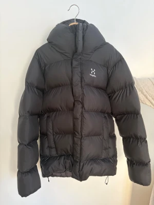Svart dunjacka från Haglöfs, S - Säljer en svart dunjacka från Haglöfs i storlek S. Jackan har en rejäl huva, puffig design och dragkedja framtill. Perfekt för kalla dagar med sin tjocka och isolerande fyllning. Diskret Haglöfs-logga på bröstet och innerfoder i svart. Passar även folk med Medium. 