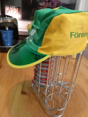 Retro keps Föreningsbanken grön/gul - Säljer en unik retro keps från Föreningsbanken i grön och gul med coolt grönt plastskärm. Kepsen har tryckt logga på sidan och är tillverkad i bomullstyg med plastdetalj framtill. Perfekt för dig som gillar vintage och vill sticka ut med färg.