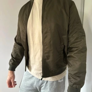 Jacka från Only & Sons - Snygg olivgrön bomberjacka från Only & Sons i storlek M. Jackan har orange foder, ribbade muddar och dragkedja framtill. Klassisk ficka på ärmen och stilren design som ger en cool vibe. Perfekt för dig som gillar streetwear och vill ha en jacka med attityd - lite nopprig i mudden men övrigt fint skick.  Modellen på bilden är 195cm.