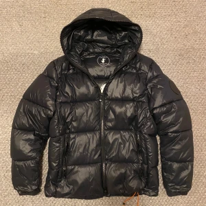 Svart pufferjacka Save The Duck  - Svart pufferjacka från Save The Duck i storlek L men skulle kunna passa M. Jackan är quiltad, har huva, dragkedja framtill och två sidofickor med dragkedja. Tillverkad i syntetmaterial och är helt animal free. Orange detaljer på dragsko och patch på ärmen. Liten skada på nedre magen med ett litet hål men de är lagat så inget som märks eller man tänker på.