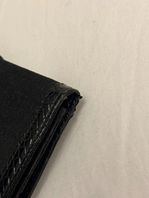 Svart plånbok från Guess med spänne - Snygg svart plånbok från Guess med ett stort metallspänne framtill och präglade detaljer. Plånboken har en stilren rektangulär form, mönstrad yta med Guess-loggor och en dragkedja på baksidan. 