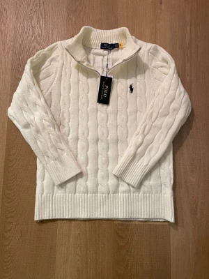Vit stickad tröja Polo Ralph Lauren - Snygg vit stickad tröja från Polo Ralph Lauren med klassisk kabelstickad design och svart broderad logga på bröstet. Tröjan har ribbad krage, muddar och nederkant samt half zip vid halsen. Perfekt för dig som gillar stilrena och tidlösa plagg.