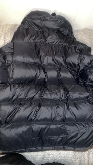 Svart pufferjacka från Burberry - Säljer en svart pufferjacka från Burberry med stor logopatch på ärmen och klassiskt rutigt foder i luvan. Jackan har dragkedja och knappar framtill, quiltad design och en riktigt schysst oversized look. Perfekt för kalla dagar och riktigt snygg till streetwear.