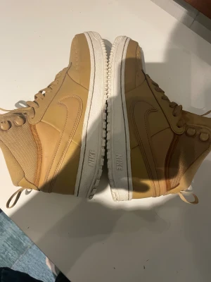 Nike Air Force 1 High Brun EUR 40 - Säljer ett par Nike Air Force 1 High i brun färg med vit sula. Skorna har snörning, högt skaft och är tillverkade i en mix av skinn och textil. Swoosh-loggan på sidan och klassisk rund tå. Perfekt för dig som gillar streetwear och vill sticka ut med snygga sneakers. använda en gång.