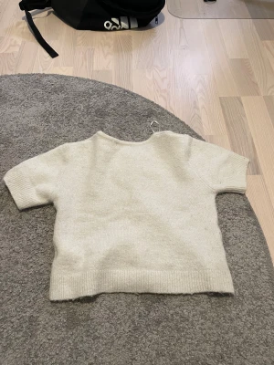 Beige stickad kofta med kort ärm - Superfin beige stickad kofta med korta ärmar och rund hals. Koftan har knappar hela vägen framtill och ribbade muddar vid ärmslut och nederkant. Snygg croppad passform som passar perfekt till jeans eller kjol.