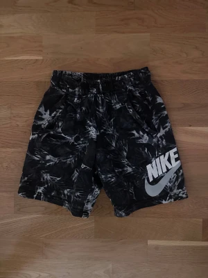 Nike svarta mönstrade shorts - Säljer ett par svarta Nike shorts med grått abstrakt mönster och stor vit Nike-logga på ena benet. De har elastisk midja och är tillverkade i mjuk bomullsmix, perfekta för chill eller träning.