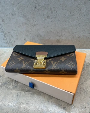 Louis Vuitton monogram plånbok - Snygg plånbok från Louis Vuitton med klassiskt monogrammönster i brunt och beige, svart lock och guldfärgad metallspänne. Perfekt storlek för kort och kontanter, med exklusiv känsla och ikonisk design.