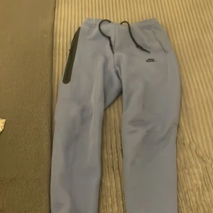 Nike ljusblå mjukisbyxor med snörning - Nike mjukisbyxor i ljusblått med svart snörning i midjan och en svart dragkedja längs ena benet. Byxorna har ribbade muddar vid bensluten och en stilren logga på framsidan. Perfekt för chill eller träning. Tvättas men tvättmaskin innan jag fraktar 