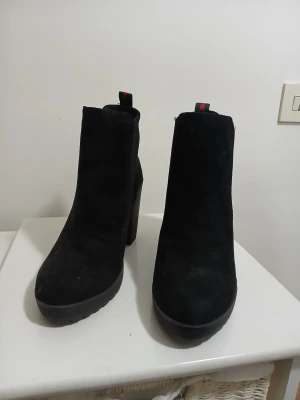 Svarta mocka stövletter med klack - Snygga svarta stövletter i mocka med hög klack och rund tå. Elastiska paneler på sidorna för enkel påtagning och en cool detalj med röd-grön rem baktill. Perfekta för dig som gillar stilrena boots med lite extra höjd.